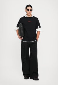 Mann mit schwarzen Sonnenbrillen, schwarzen übergroßen Hosen, schwarzem T-Shirt über einem weißen Hemd, der eine schwarze Leder-Clutch-Tasche hält.