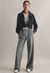 Granatowa kurtka na zamek z białymi paskami, zestawiona z szarymi spodniami z wysokim stanem, plisami i sznurkiem w pasie. Dopełnieniem są białe trampki.