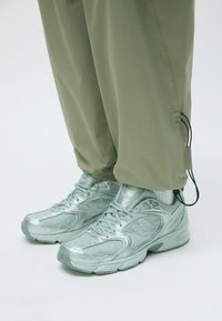 Metaliczne srebrne sneakersy z teksturowanymi detalami i zakrzywionym kształtem, zestawione z luźnymi oliwkowymi spodniami z troczkiem przy dolnej krawędzi.