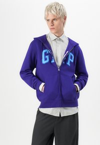 Jonge man met kort blond haar, gekleed in een paarse GAP-ritsjas over een witte gestreepte blouse en donkergekleurde broek, met zijn handen in zijn zakken.