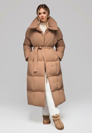 Femme blonde debout, portant un long manteau beige matelassé avec une ceinture, un pantalon blanc et des bottes d'hiver en fourrure beige sur fond gris.