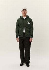 Mladý muž stojí, oblečený v tmavozelenej mikine na zips s logom LD, čiernych nohaviciach, bielom tričku, čiernych topánkach a svetlo béžovej baseballovej čiapke.