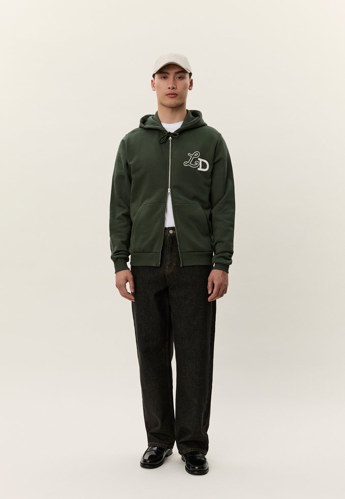 Mladý muž stojí, oblečený v tmavozelenej mikine na zips s logom LD, čiernych nohaviciach, bielom tričku, čiernych topánkach a svetlo béžovej baseballovej čiapke.