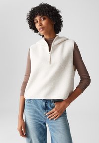 Weißer Fleece-sansärmeligen Kapuzenpullover mit hohem Kragen und Frontreißverschluss, kombiniert mit einem braunen Langarmoberteil und hellblauen Jeans.