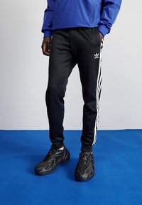 Černé atletické kalhoty s bílými pruhy na bocích a logem Adidas. Kombinované s černými texturovanými teniskami, vše na modrém pozadí.