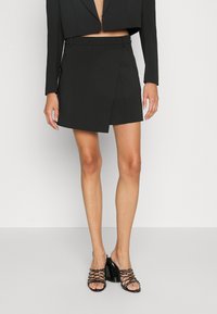 Svart asymmetrisk omlottskjolt matchad med svarta sandaler med klack och remmar på en stående person framför en neutral bakgrund.