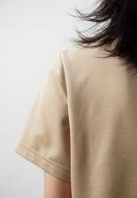 Béžová košile s krátkým rukávem, která má texturovanou látku, hladké švy a volný střih, viděná zezadu s částečně viditelnými vlasy.