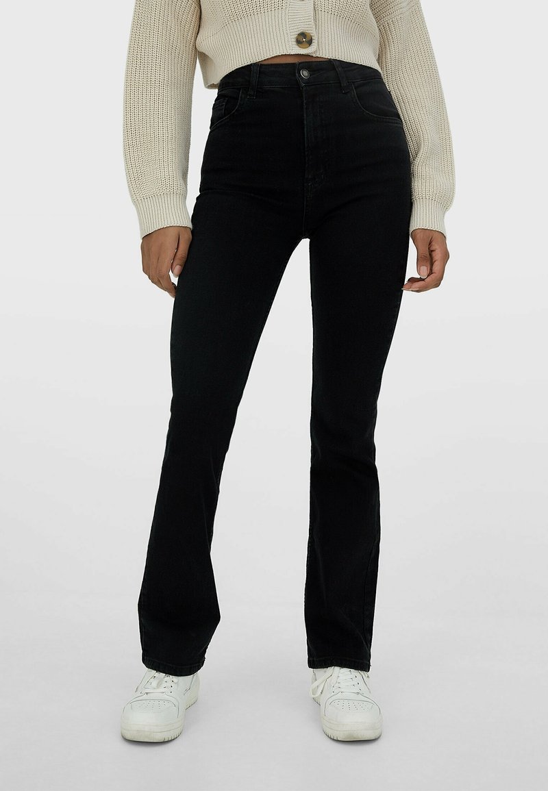 Femme portant un jean noir évasé taille haute, des baskets blanches et un cardigan court en tricot beige avec un gros bouton.