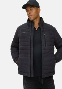 Sort pufferjakke med vandrette quiltninger, høj krave, lynlås-lomme på brystet og sort sweater under, parret med en kasket.