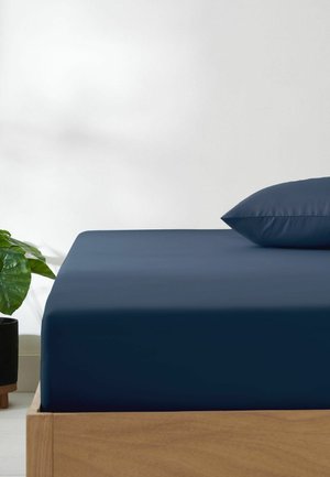 Holzrahmen für ein Bett mit marineblauem Spannbettlaken und passendem Kissen, neben einem schwarzen Topf mit großen grünen Blättern an einer weißen Wand.