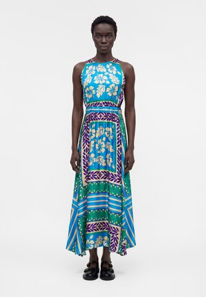 Γυναίκα στέκεται από εμπρός, φορώντας αμάνικο, μακρύ φόρεμα με σχέδια σε μπλε, πράσινο, μωβ και μπεζ με ανθικά και γεωμετρικά μοτίβα.