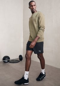 Olívazöld pulóver bordázott mandzsettával, fekete sortiás rövidnadrággal párosítva, amelyen fehér logó található. A megjelenést fekete sportcipő és fehér zokni teszi teljessé.