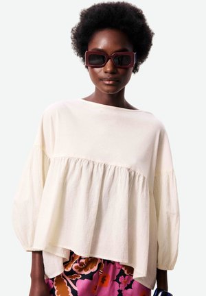 Femme avec coiffure afro portant un chemisier ample beige clair, de grandes lunettes de soleil rectangulaires bordeaux et une jupe fleurie rose et marron.