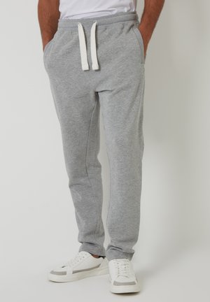 Man draagt grijze joggingbroek met witte trekkoorden en witte sneakers, staand met handen in de zakken.
