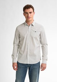 Camicia bianca con pois neri, maniche lunghe e tasca sul lato sinistro del petto. Indossata con jeans blu.
