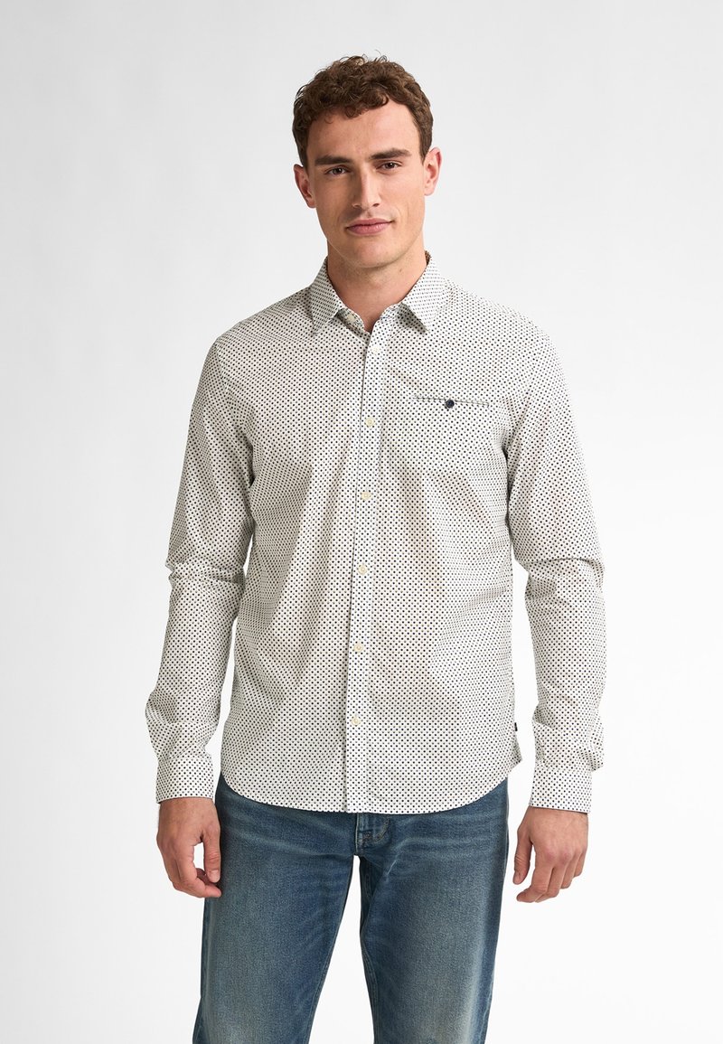 Camicia bianca con pois neri, maniche lunghe e tasca sul lato sinistro del petto. Indossata con jeans blu.