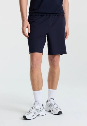 Mand iført sorte atletikshorts, hvide crew-sokker og hvide New Balance løbesko, stående på en ensfarvet lys baggrund.