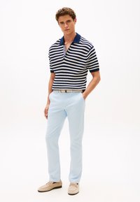 Πουκάμισο πόλο με ρίγες σε ναυτικό μπλε και άσπρο, κοντά μανίκια, με γιακά· συνδυασμένο με ανοιχτό μπλε παντελόνι και μπεζ λοφρέρ, σε απλό φόντο.