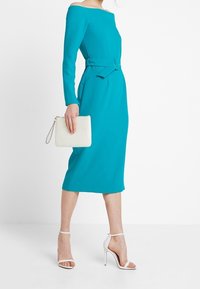 Tyrkysové šaty s odhalenými rameny a dlouhými rukávy, páskem v pase a délka ke kolenům; kombinované s bílou kabelkou a bílými sandály na vysokém podpatku.