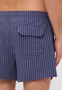 Marineblå stripete badeshorts laget av lett materiale, med en baklomme med klaff og to trykknapper for sikker lukking.