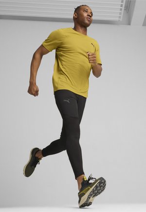 Dzeltena sporta krekls ar īsām piedurknēm, apvienots ar melnām legingām un melniem skriešanas apaviem ar teksturētu zoli, dinamiskā skriešanas pozā.