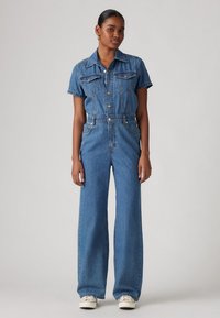 Jeans overall med krage, korte ermer, frontknapper, vide ben og brystlommer i middels blå vask.