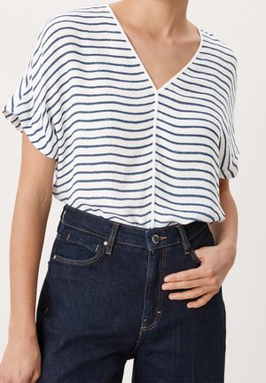 Frau trägt eine lockere weiße V-Ausschnitt-Bluse mit marineblauen horizontalen Streifen, die in dunkelblaue High-Waist-Jeans gesteckt ist. Eine Hand ruht auf der Hüfte.