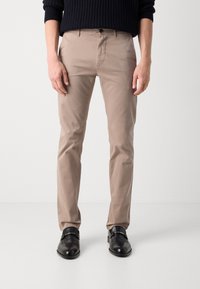 Pantalons en coton beige avec une coupe ajustée, présentant une fermeture à bouton, des poches latérales et une texture lisse. Portés avec des chaussures en cuir noires.