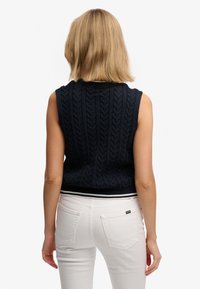 Donkerblauwe mouwloze gebreide top met kabelpatroon en geribbelde zoom, gecombineerd met witte, aansluitende broek met achterzakken.