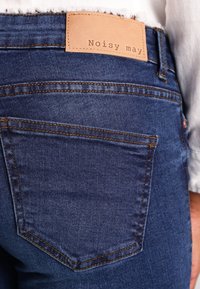 Mörkblå denimjeans med en slät textur. Funktioner inkluderar en läderpatch med texten "Noisy may" och synlig sömnad på bakfickorna.