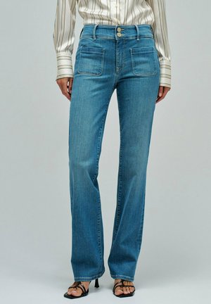 Mujer con jeans de mezclilla azul de cintura alta y bolsillos delanteros parche, combinados con sandalias negras de tacón con tiras y una camiseta de manga larga a rayas.