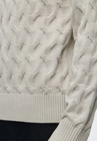 Pull en maille torsadée beige clair avec jupe côtelée et poignets aux manches, porté par une personne portant un pantalon noir.