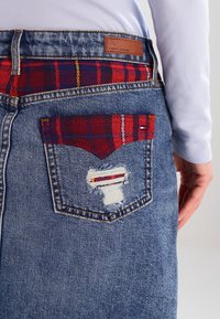 Denim-hame sinisellä pesulla, repaleisella taskuyksityiskohdalla ja punaisella ruudullisella vyötärönauhalla. Takana on ruskea nahkakangasmerkki.