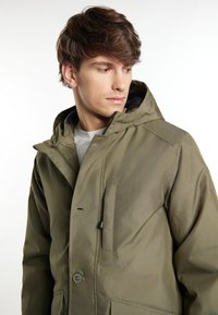 Giacca verde oliva con cappuccio, caratterizzata da una chiusura con zip, accenti a bottoni e tasche laterali. Realizzata in materiale sintetico strutturato.