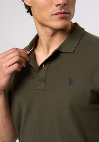 Oliven grønn polo skjorte med ribbet krage, med en marineblå brodert logo på venstre bryst og en kontrasterende detalj på den indre kragen.