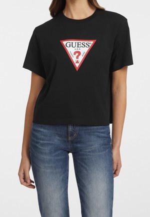 Kobieta ubrana w czarny krótki t-shirt z czerwonym trójkątnym logo Guess oraz niebieskie, wyblakłe dżinsy na białym tle.