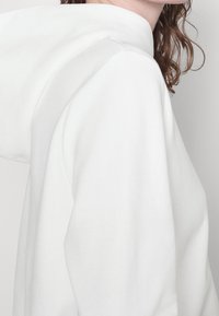 Sudadera blanca con capucha que presenta un bolsillo tipo canguro, puños acanalados y una textura suave y lisa. El diseño es simple y minimalista.