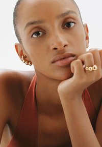 La joyería de oro incluye un pendiente de cuentas y un anillo con acentos circulares. El modelo tiene la piel suave y lleva una blusa sin mangas de color óxido.