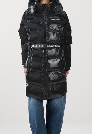 Błyszcząca czarna pikowana kurtka z kapturem i przednim zamkiem błyskawicznym, z paskiem w talii z napisem "LAGERFELD", noszona na niebieskich dżinsach.