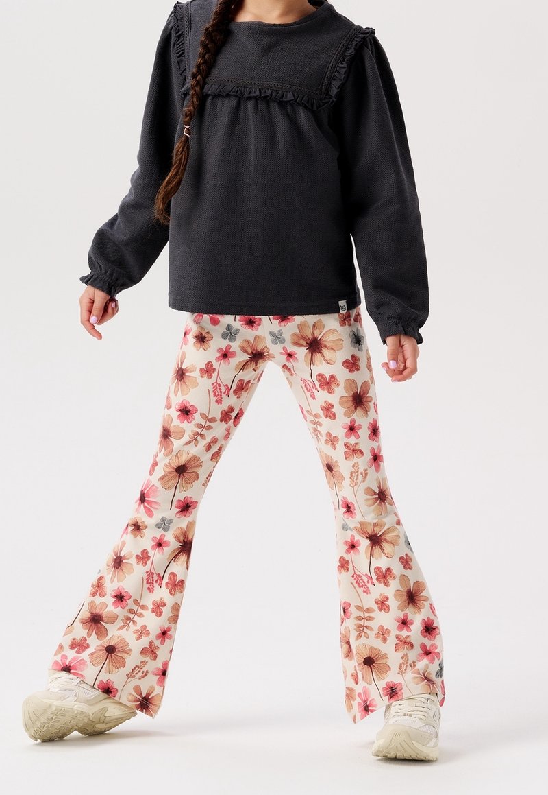 Enfant portant un haut noir à manches longues avec des volants et un pantalon évasé crème à fleurs avec des baskets blanches, debout les jambes écartées.