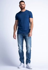 Niebieska bawełniana koszulka z okrągłym dekoltem, w połączeniu z jasnoniebieskimi obcisłymi dżinsami. Mężczyzna stoi w białych trampkach na jednolitym tle.