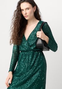 Abito di paillettes verdi lucidi con maniche lunghe e scollatura a V profonda. La vita è stretta e una borsa nera è tenuta con una mano.
