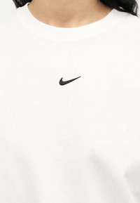 Κοντινό πλάνο ενός ατόμου που φοράει λευκό μπλουζάκι με στρογγυλή λαιμόκοψη και ένα μικρό μαύρο λογότυπο Nike Swoosh στο κέντρο κάτω από το γιακά.