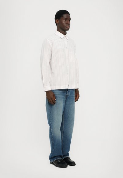 Chemise à rayures blanche avec des lignes rouges et bleues, associée à un jean droit bleu clair et des chaussures noires. Look propre et décontracté.
