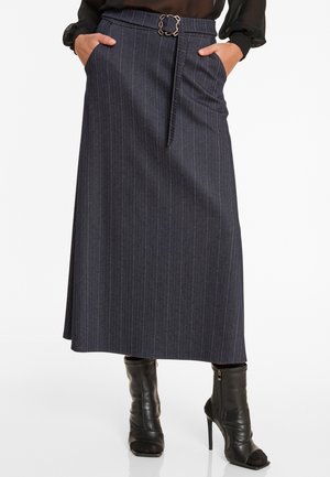 Vrouw in een marineblauwe pinstripe midi-jurk met een kettingriem en zwarte hakken enkellaarsjes, handen in de zakken.