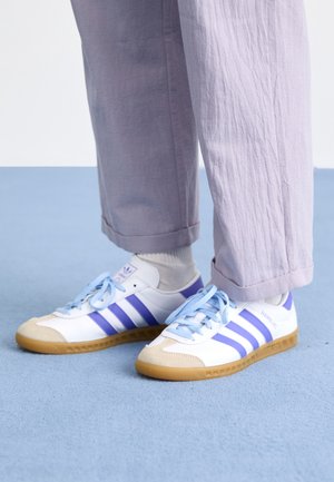 Voeten met witte sneakers met blauwe strepen en lichtblauwe veters, gecombineerd met lichtpaarse broek en witte sokken op een blauwe vloer.