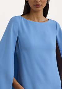 Blå topp med cape som är tillverkad av ett slätt tyg. Rund halsringning, med flödande cape-liknande ärmar som sträcker sig från axlarna och skapar en smidig silhuett.