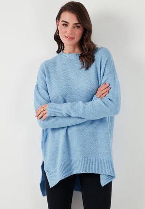 Maglione lavorato a maglia di colore blu chiaro, con scollatura rotonda, spalle scese e orlo a coste. Presenta fessure laterali per facilitare il movimento.