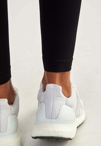 White athletic shoes with textured mesh upper, a soft heel counter, and contrasting black accents, worn with black textured leggings.
