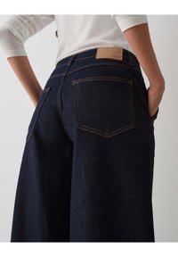 Dark blue denim jeans with a wide leg, rear pockets, and a tan leather label at the waistband. Features contrasting orange stitching.